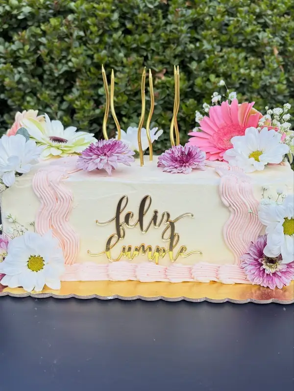 Torta rectangular con flores naturales rosadas y blancas y letras doradas «Feliz cumple»